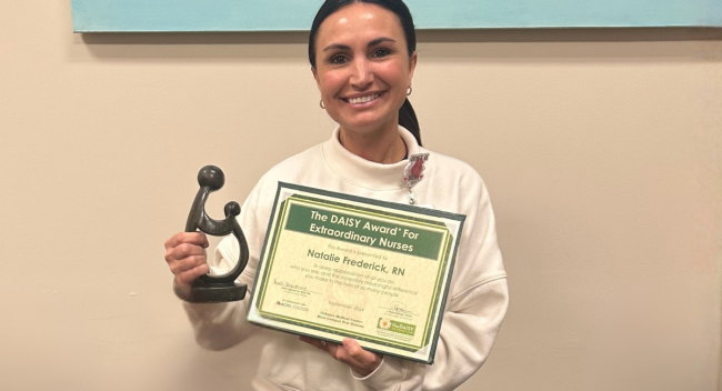 RN to BSN online student smiles with her DAISY Award, wearing a white sweater and blue scrub pants.